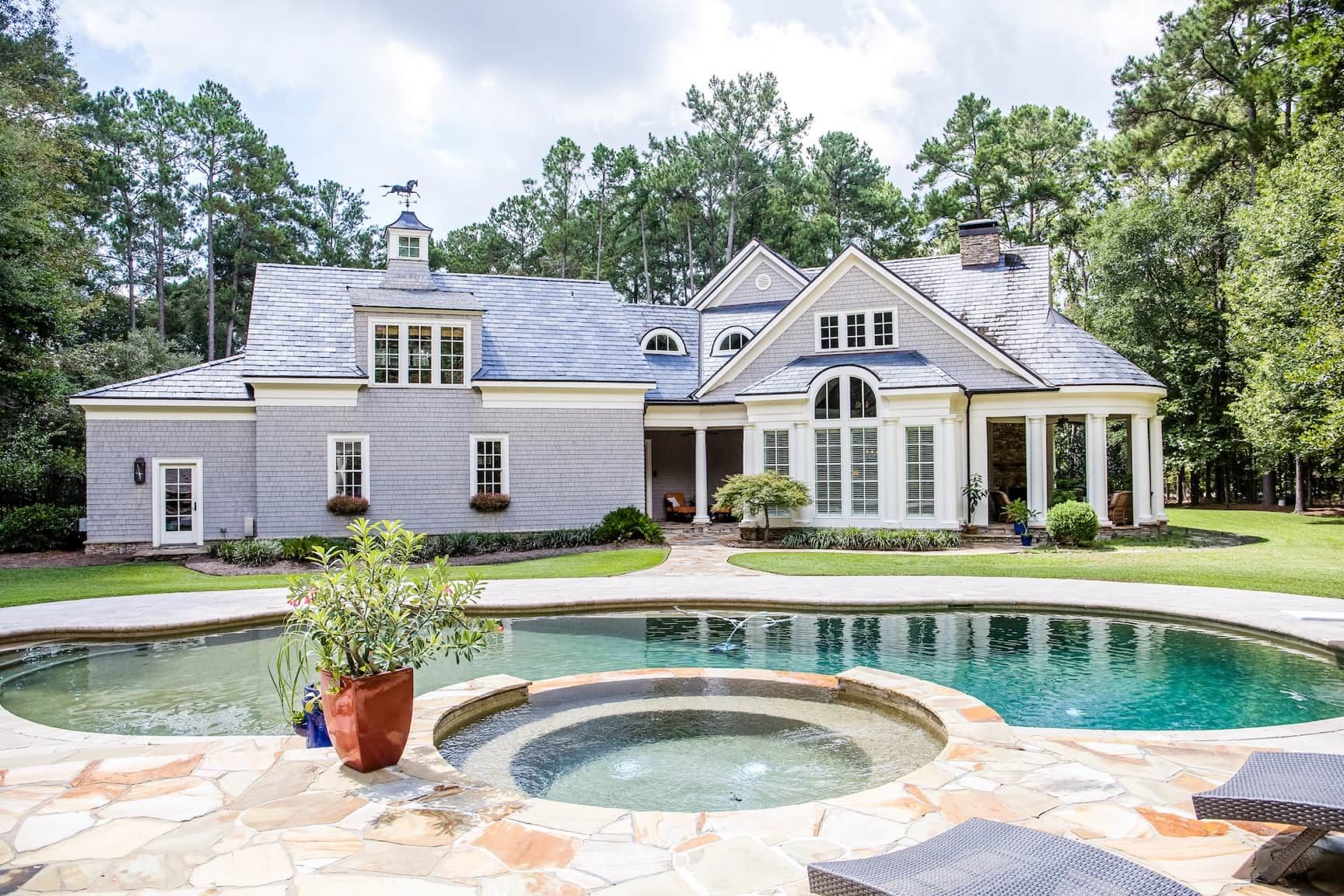 A freeform pool and round spa on a flagstone deck with a wide lawn behind a shingle-sided home