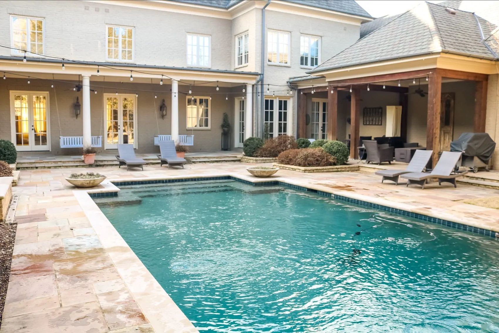 A rectangular pool with tile trim, cushioned loungers, and a covered patio behind a two-story brick home