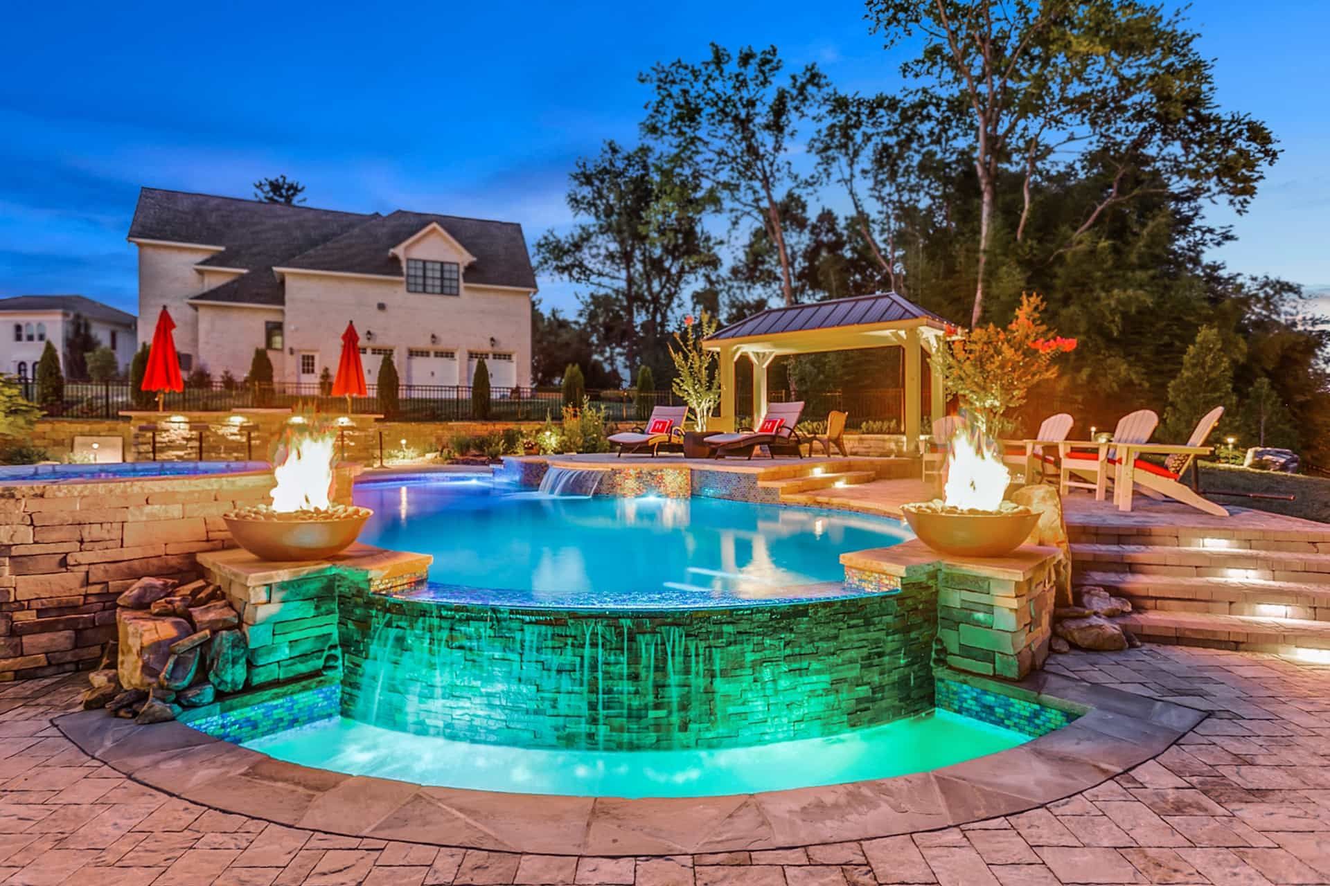 A freeform pool and glass-tile spa with fire bowls and a poolside gazebo lit at dusk